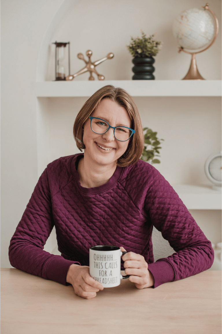 A woman wearing a burgundy sweater is sitting at a desk holding a mug. The mug says "this calls for a spreadsheet". The woman is smiling and has a blonde bob haircut, and blue reading glasses